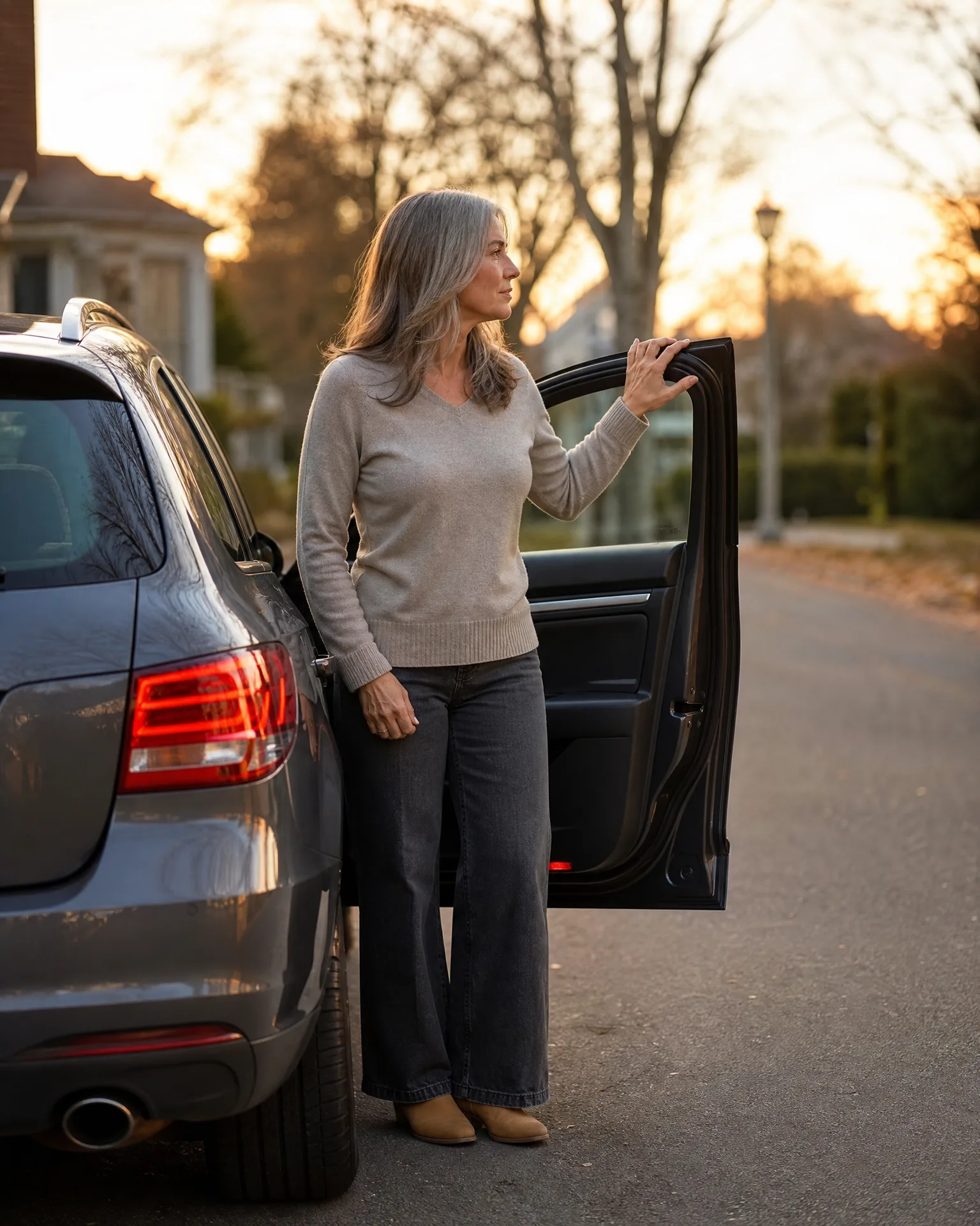 A woman with long gray hair in a beige sweater and dark wide-leg jeans stands beside a dark SUV on a street at sunset, holding the open front passenger door.