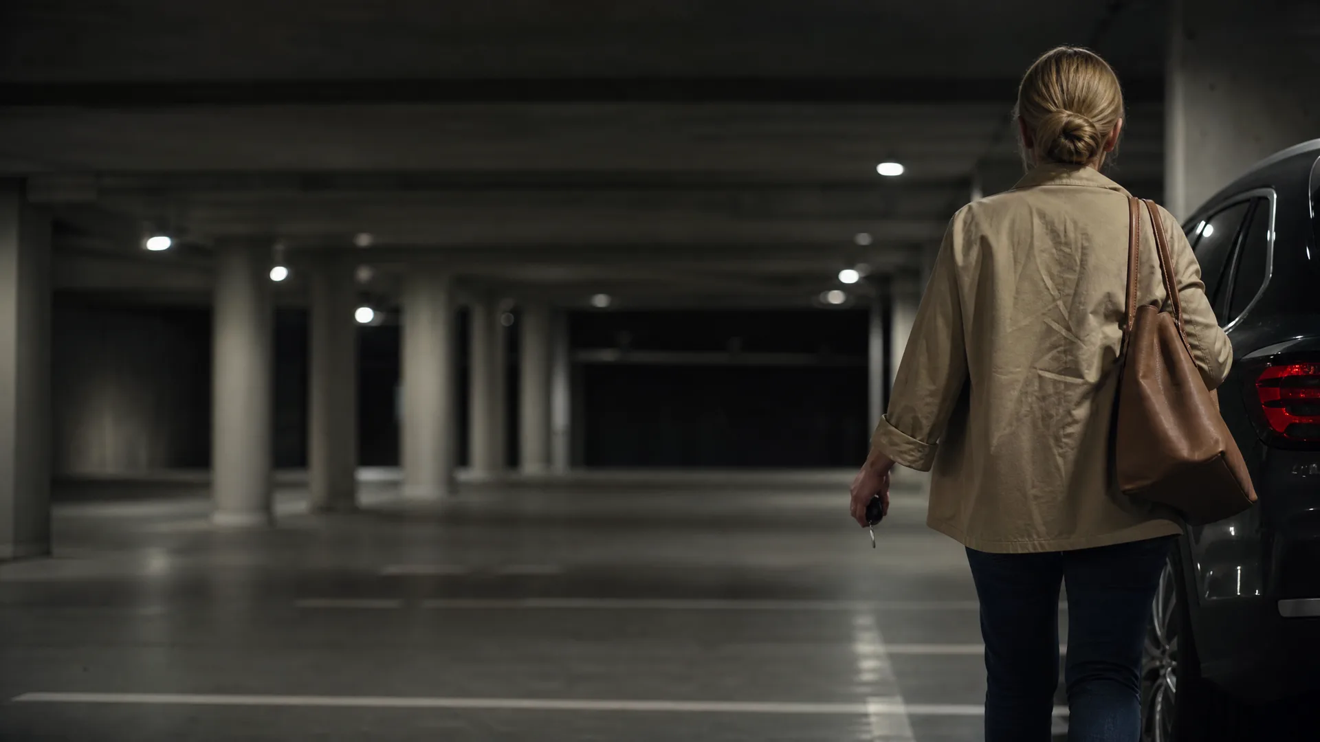 A woman seen from behind in a beige jacket, jeans and brown shoulder bag holds car keys while walking toward a dark car in a dimly lit underground parking garage with concrete columns.