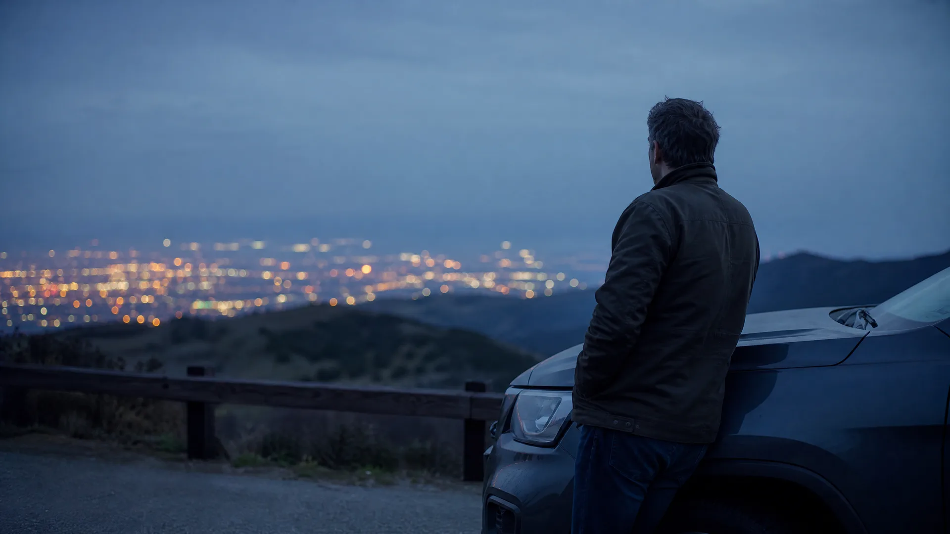 A man in a dark jacket stands leaning against the front of a dark SUV at a roadside overlook at dusk, facing away toward distant city lights spread across a valley below hills.