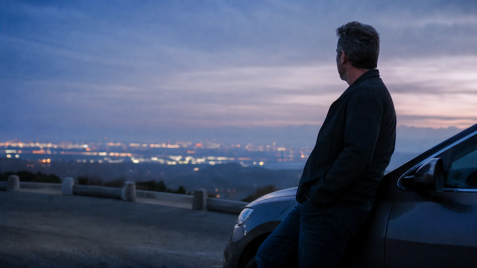 A man in a dark jacket leans against the front of a car on a hilltop overlook at dusk, looking out over distant city lights under a cloudy purple sky.