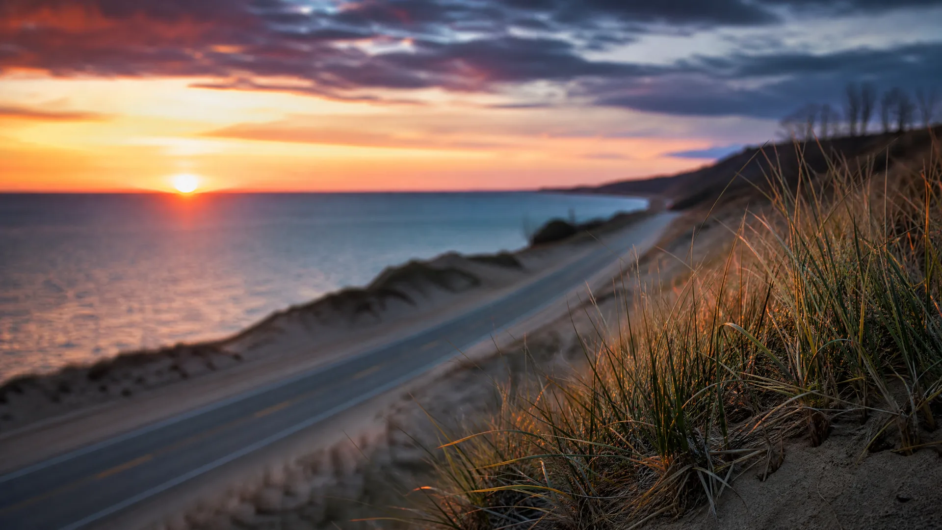 A two-lane road curves along a grassy sandy bluff above a large body of water at sunset, with dune grass in sharp focus in the foreground and orange and purple clouds overhead.