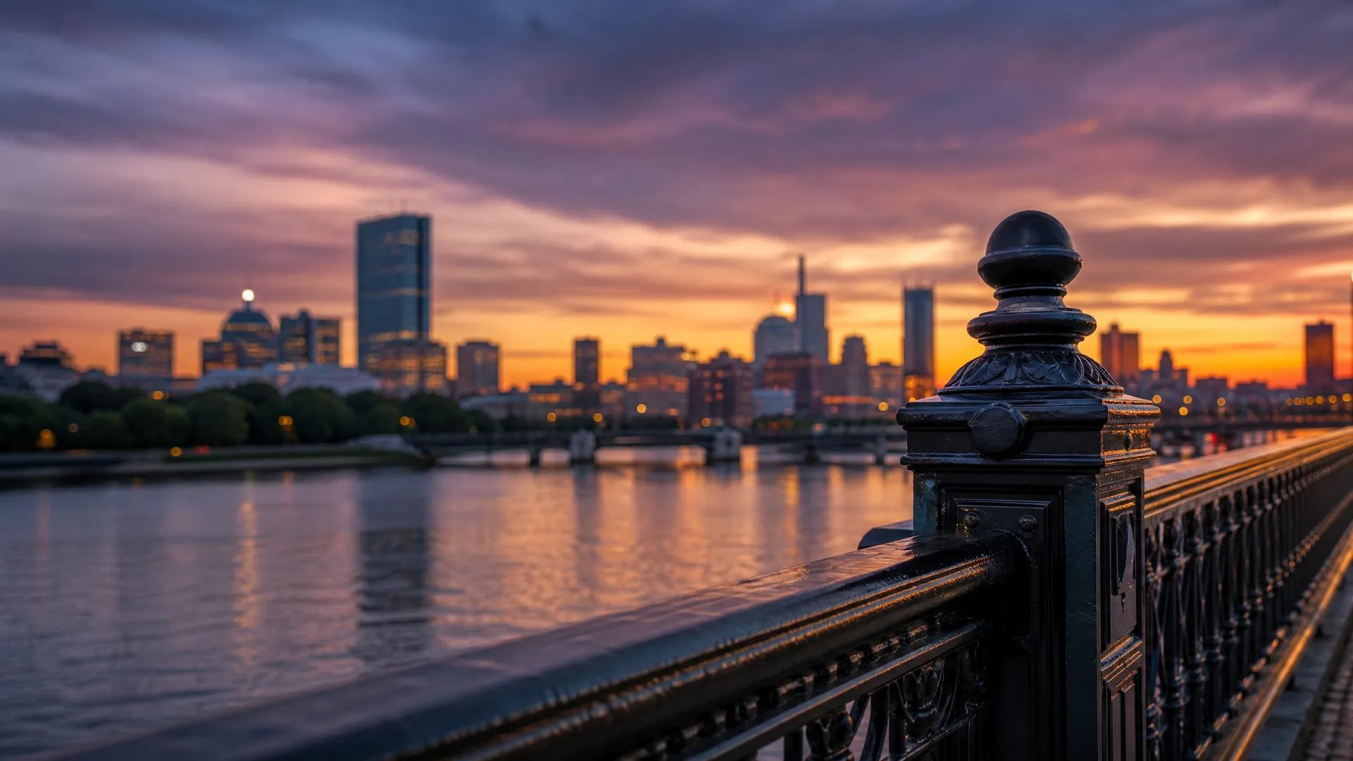 A dark ornate iron bridge railing with a ball-topped post in the foreground overlooks a river and a blurred city skyline at sunset under orange and purple clouds.